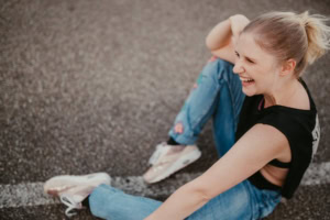 Lächelnde junge Frau auf Asphaltboden - photoart hübner business Junge Frau sitzt lachend auf Asphalt, trägt sportliche Kleidung, ideal für Business- und Karrierefot.