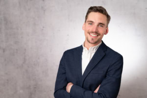 Business-Mann in Anzug, lächelnd, vor neutralem Hintergrund.