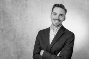 Business-Porträt eines Mannes in Anzug, lächelnd und selbstbewusst, vor neutralem Hintergrund.