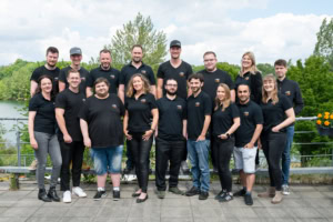 Teamfoto eines Unternehmens mit 16 Mitarbeitenden im Freien, alle in schwarzen T-Shirts. - photoart hübner business Mitarbeitende im Teamfoto für Business- und Unternehmensfotografie.
