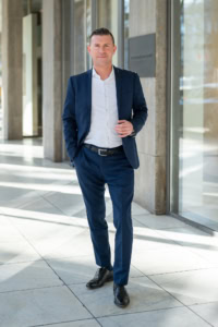 Selbstbewusster Mann in Anzug vor moderner Firmenfassade - photoart hübner business Geschäftsmann in Businesskleidung vor Bürogebäude, professionell und selbstsicher.