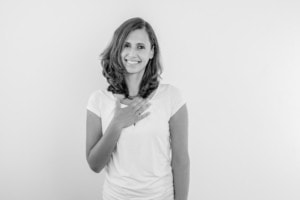 Businessfotografie Portrait einer Frau vor weißem Hintergrund - photoart hübner business Frau in Businesskleidung, lächelnd, mit Hand auf der Brust, professionell und freundlich.