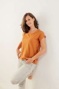 Businessfotografie Portrait einer jungen Frau in casual Kleidung - photoart hübner business Junge Frau in orangefarbenem T-Shirt und weißen Hosen, lächelnd, vor weißem Hintergrund.