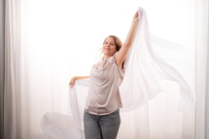 Fröhliche Frau mit weißen Tüchern, die Freiheit und Leichtigkeit symbolisieren - photoart hübner business Frau mit weißen Tüchern, die Freiheit und Leichtigkeit in einem hellen Raum darstellen.