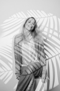 Moderne Businessfotografie mit natürlichem Lächeln - photoart hübner business Frau in Businesskleidung sitzt vor einer weißen Wand mit Schatten von Palmenblättern.