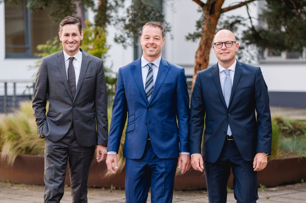 Erfolgreiche Geschäftsmänner im Business-Anzug vor Bürogebäude - photoart hübner business Drei Männer in Anzügen stehen im Freien vor einem Bürogebäude.