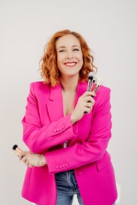Businessfotografie mit Frau in pinkem Blazer und Make-up-Tools - photoart hübner business Frau in pinkem Blazer hält Make-up-Pinsel, lächelt vor weißem Hintergrund.