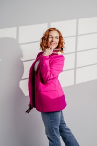 Businessfotografie mit Frau in pinkem Blazer und Posaune - photoart hübner business Fröhliche Frau in Businessoutfit mit Posaune vor Fenster, professionell fotografiert.