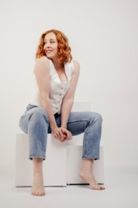Businessfotografie Portrait mit rotem Haar und natürlichem Look - photoart hübner business Frau mit roten Haaren in Businesskleidung, sitzt entspannt auf weißen Quadern.