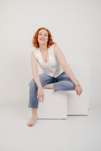 Selbstbewusste Frau in Casual-Outfit vor weißem Hintergrund - photoart hübner business Frau sitzt entspannt auf weißen Kuben, lächelt freundlich, ideal für Personal Branding und Karrieref.