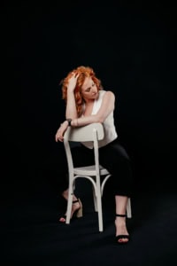 Businessfotografie mit Model und Stuhl - photoart hübner business Frau mit roten Haaren posiert auf einem Stuhl vor schwarzem Hintergrund.