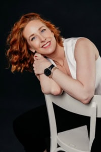 Porträt einer jungen Frau mit roten Locken, lächelnd, in Businesskleidung, auf schwarzem Hintergrund. - photoart hübner business Porträt einer professionellen Frau für Business- und Personal Branding Fotos.