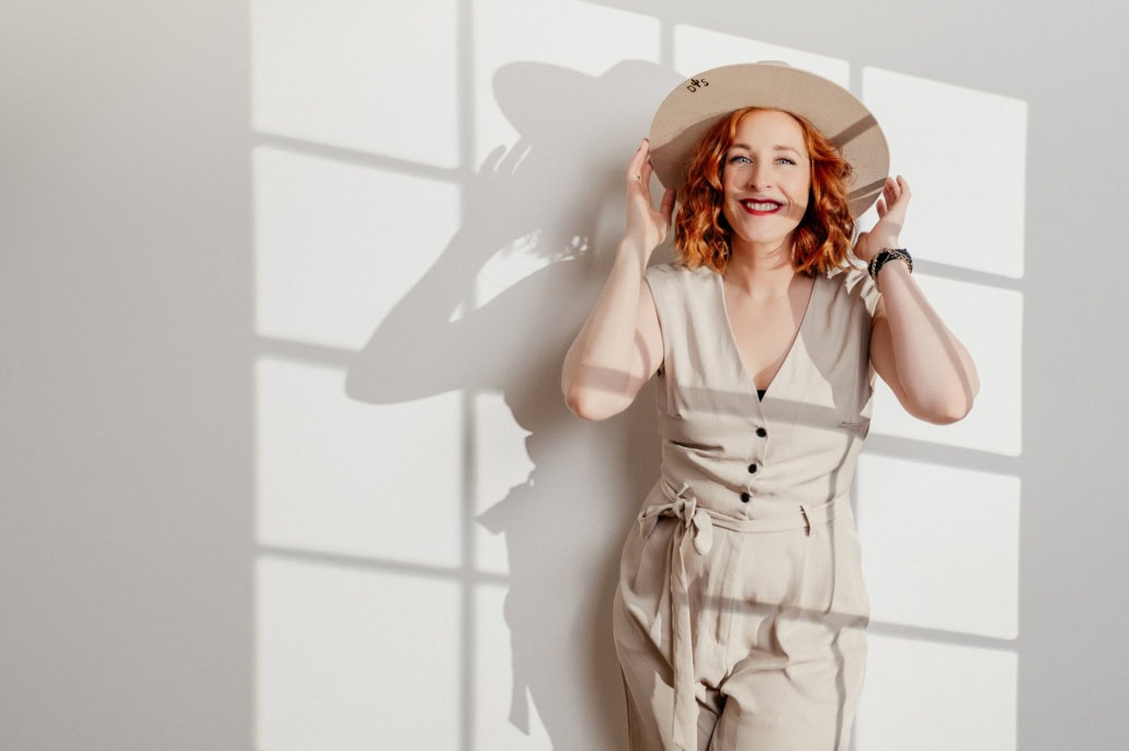 Moderne Portraitfotografie mit natürlichem Licht und kreativer Beleuchtung - photoart hübner business Fröhliche Frau in beige Outfit mit Hut vor Schattenmuster an Wand.
