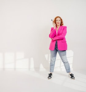 Businessfotografie mit Frau in pinkem Blazer und Jeans, professionell und modern. - photoart hübner business Frau in Businesskleidung posiert selbstbewusst mit Smartphone vor weißem Hintergrund.